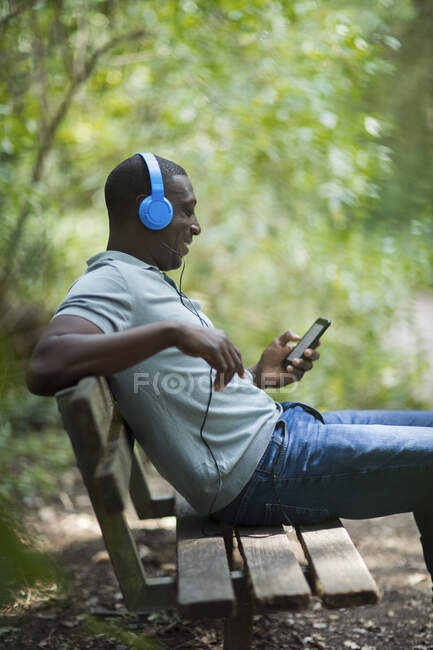 Homem ouvindo música com fones de ouvido e telefone inteligente no banco do parque — Fotografia de Stock