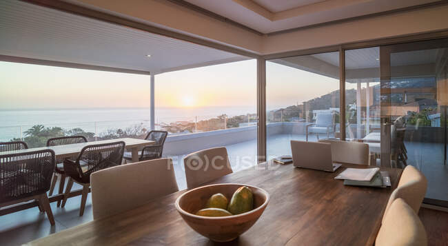 Ordenador portátil en mesa de comedor de lujo con vistas panorámicas al océano al atardecer - foto de stock