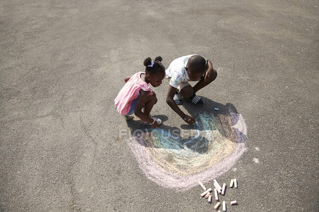 Hermano y hermana dibujando arco iris con tiza de acera en el pavimento - foto de stock