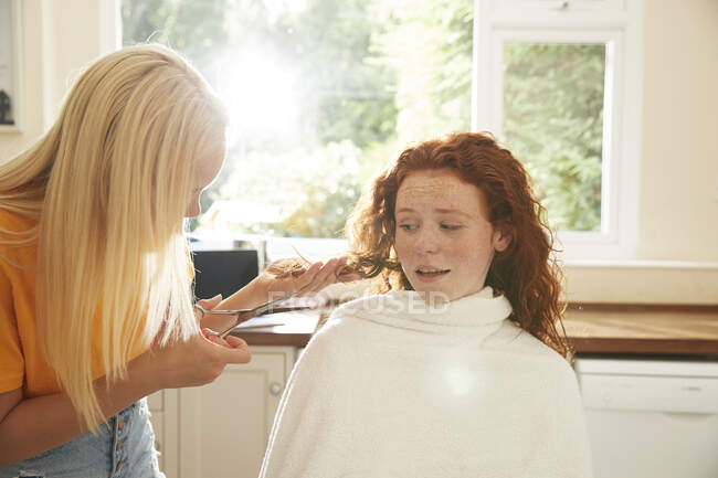 Teenage girl cutting hair for worried friend in sunny kitchen — Stock Photo