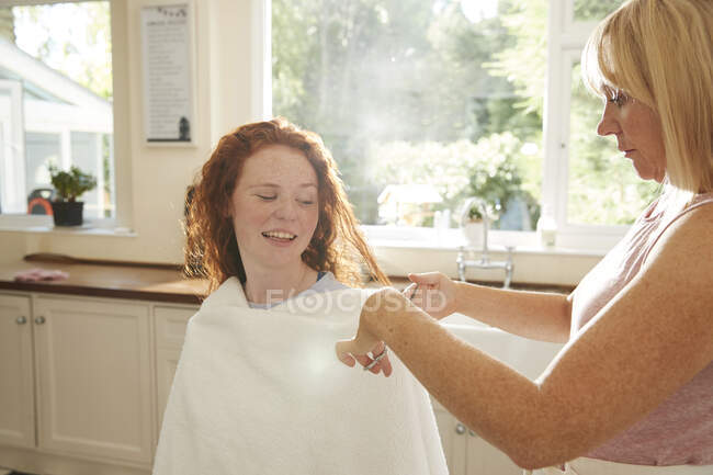 Mãe corte de cabelo para a filha pré-adolescente na cozinha ensolarada — Fotografia de Stock