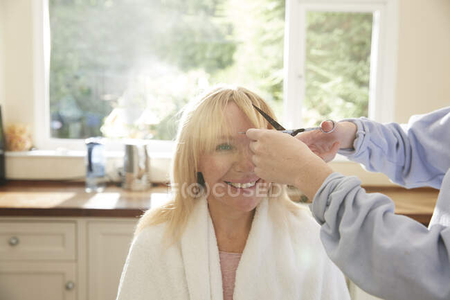Daughter cutting bangs for happy mother in sunny kitchen — Stock Photo