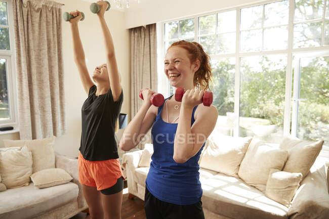 Felices amigas preadolescentes haciendo ejercicio con pesas en la sala de estar - foto de stock