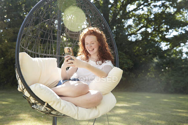 Joyeux preteen fille en utilisant le téléphone intelligent dans la chaise oscillante dans la cour ensoleillée — Photo de stock