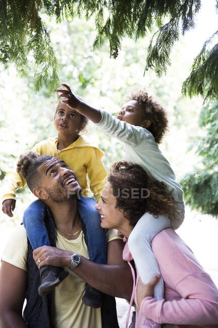 Pais felizes que transportam filhas em ombros abaixo de árvores no parque — Fotografia de Stock