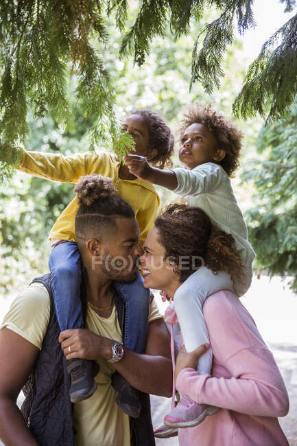 Affectionate couple carrying daughters on shoulders below park trees — Stock Photo