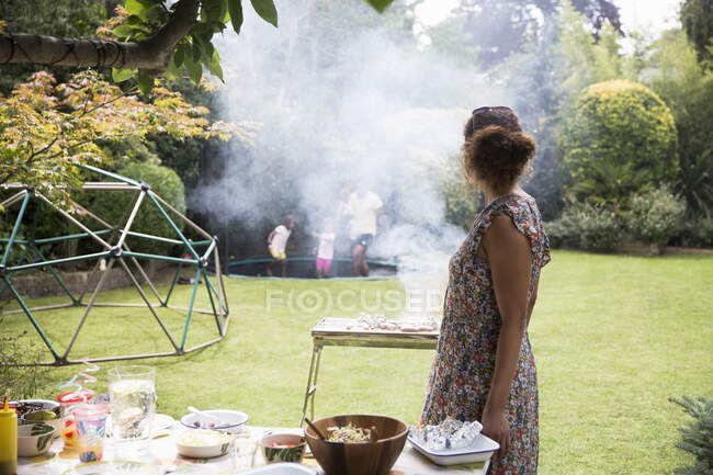 Donna barbecue e guardare la famiglia giocare sul trampolino da giardino — Foto stock