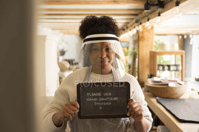 Retrato femenino dueño de la tienda en escudo facial sosteniendo la mano desinfectante signo - foto de stock