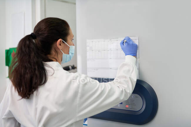 Cientista feminina na máscara facial marcação cronograma na parede em laboratório — Fotografia de Stock
