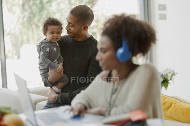 Father holding baby daughter behind mother working at laptop — Stock Photo