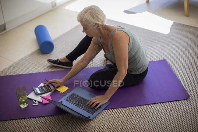 Старшая женщина оплачивает счета и упражнения на ноутбуке на коврике для йоги — стоковое фото