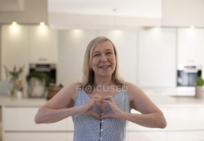 Portrait beautiful happy senior woman gesturing heart shape in kitchen — Stock Photo