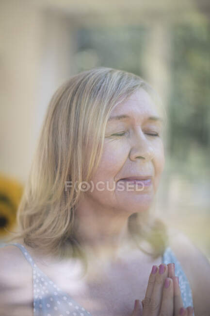 Serene senior woman praying with hands clasped at window — Stock Photo