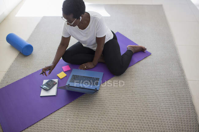 Зрелая женщина оплачивает счета и работает за ноутбуком на коврике для йоги — стоковое фото