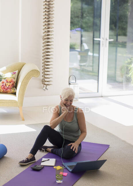 Donna anziana con cuffie che lavora al computer portatile sul tappetino yoga a casa — Foto stock