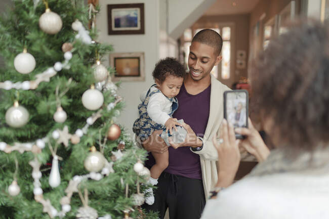 Woman photographing husband and baby daughter at Christmas tree — Stock Photo