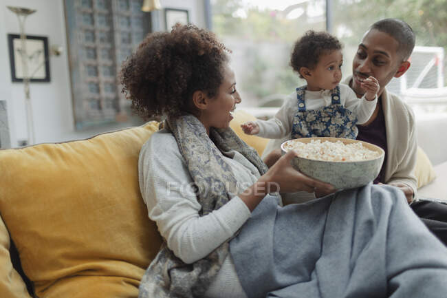 Pais felizes e bebê filha assistindo TV com pipoca no sofá — Fotografia de Stock