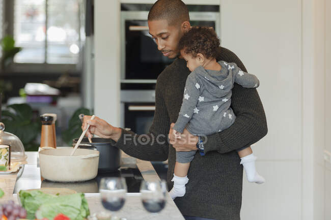 Father holding baby daughter and cooking dinner at kitchen stove — Stock Photo