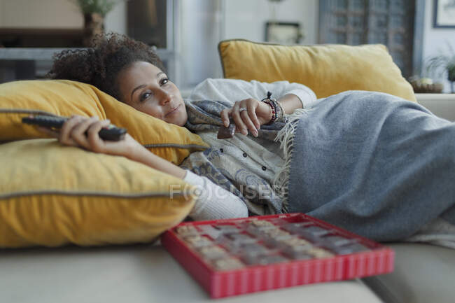 Mulher acolhedora comer chocolates e assistir TV no sofá da sala de estar — Fotografia de Stock