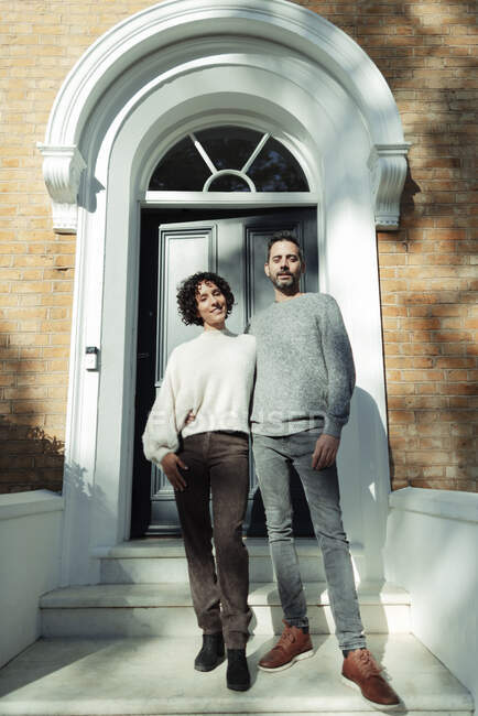 Портрет уверенной пары у арочной входной двери на солнечном крыльце дома — стоковое фото