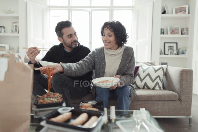 Feliz casal desfrutando de comida de takeout com pauzinhos no sofá da sala de estar — Fotografia de Stock