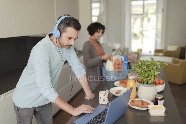 Человек с наушниками работает из дома за ноутбуком на кухне — стоковое фото