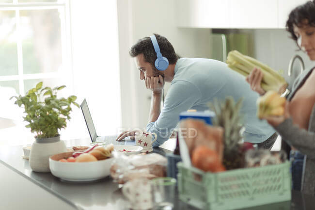Чоловік з навушниками, що працюють з дому на ноутбуці на кухні — стокове фото