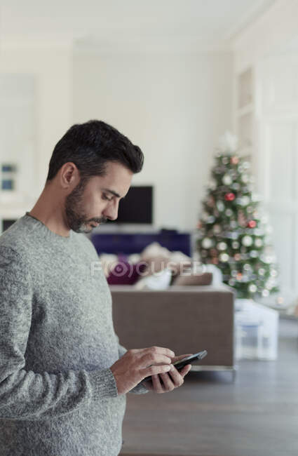 Homem usando telefone inteligente na sala de estar com árvore de Natal — Fotografia de Stock