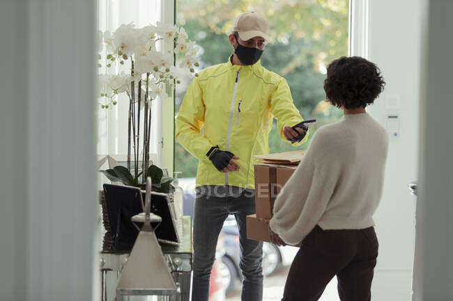 Mensajero en máscara facial entrega de paquetes a la mujer en casa - foto de stock