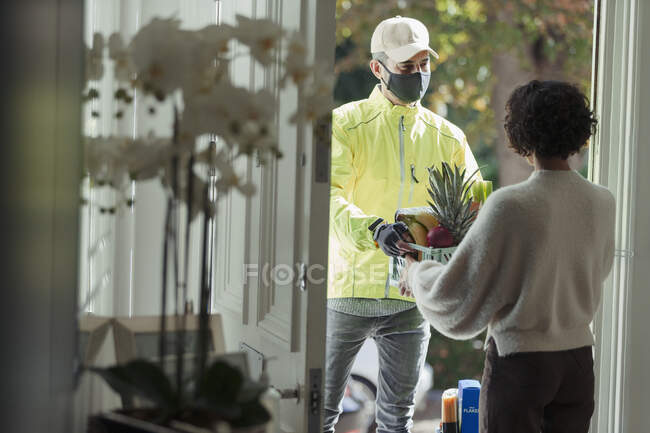 Женщина, получающая доставку продуктов от курьера в маске на двери — стоковое фото