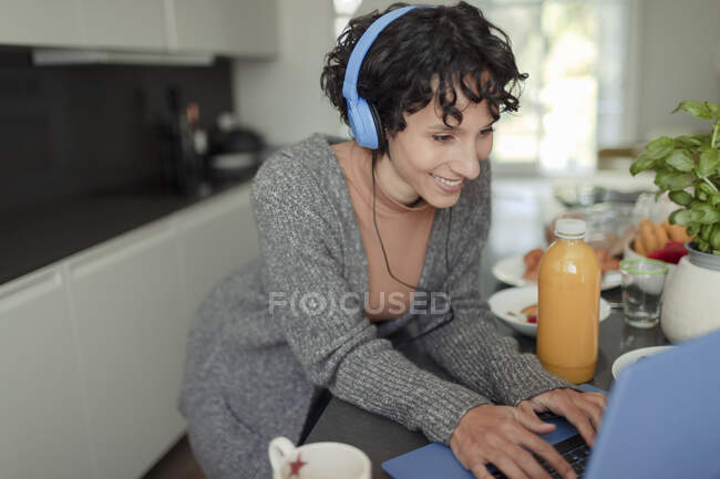Щаслива жінка з навушниками, що працюють з дому на ноутбуці на кухні — стокове фото
