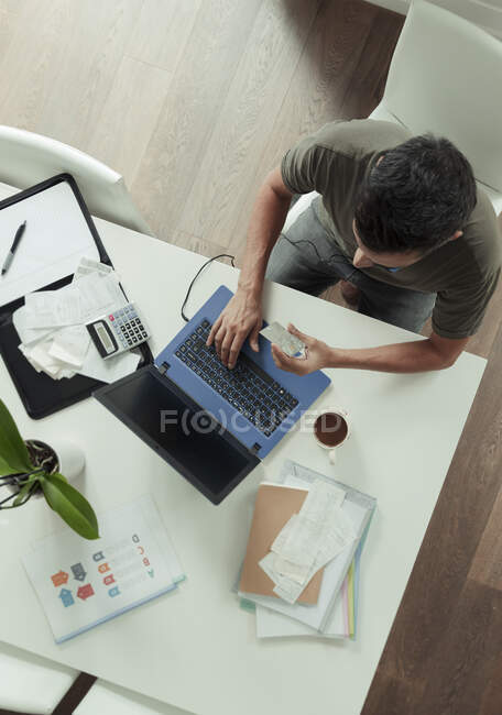 Человек с кредитной картой оплачивает счета за ноутбук на обеденном столе — стоковое фото
