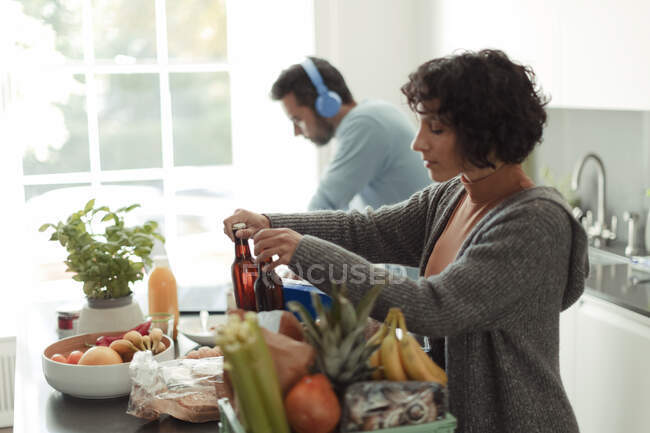 Femme déchargement épicerie dans la cuisine tandis que le mari travaille à l'ordinateur portable — Photo de stock
