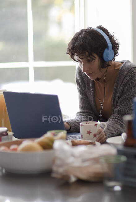 Femme avec écouteurs travaillant de la maison à l'ordinateur portable dans la cuisine — Photo de stock