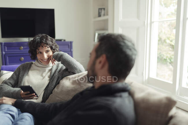 Casal feliz com telefone inteligente relaxante e falando no sofá da sala de estar — Fotografia de Stock