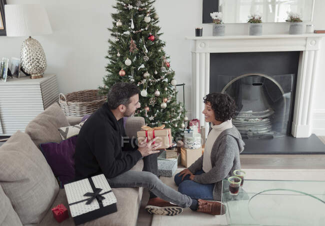 Husband giving Christmas gift to wife by tree in living room — Stock Photo