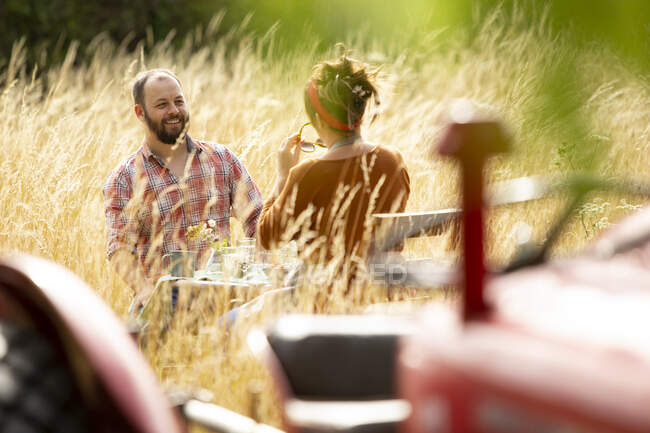 Happy couple at table behind tractor in sunny summer tall grass — Stock Photo