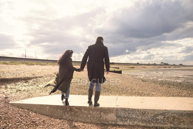Пара в зимних пальто, держащихся за руки, гуляющих по солнечному пляжу — стоковое фото
