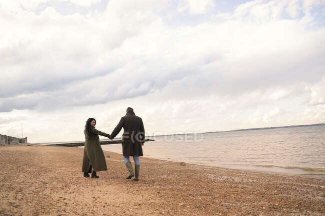 Pareja en abrigos de invierno tomados de la mano caminando en la playa del océano - foto de stock