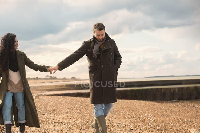 Пара в зимних пальто, держащихся за руки, гуляющих по солнечному пляжу — стоковое фото