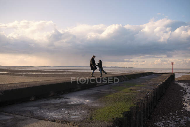 Happy couple in winter coats on sunny ocean beach jetty — Stock Photo