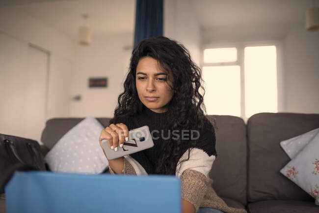 Жінка працює вдома з розумним телефоном і ноутбуком на дивані. — стокове фото