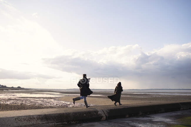 Casal em casacos de inverno correndo no ensolarado oceano praia molhe — Fotografia de Stock