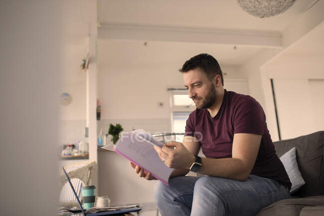 Человек с бумагами работает из дома за ноутбуком на диване — стоковое фото