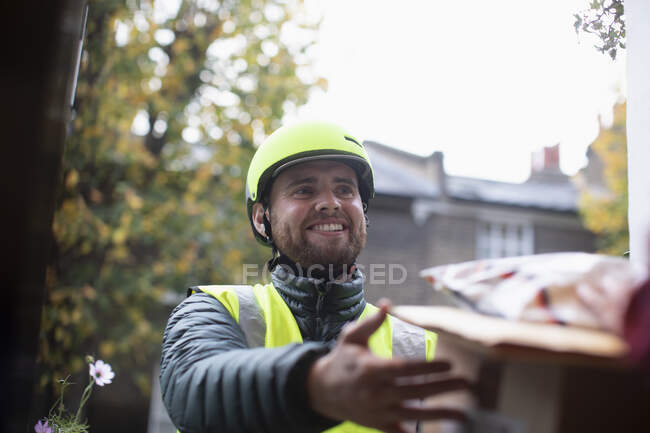 Щасливий дружній чоловічий кур'єр в шоломі робить доставку біля вхідних дверей — стокове фото