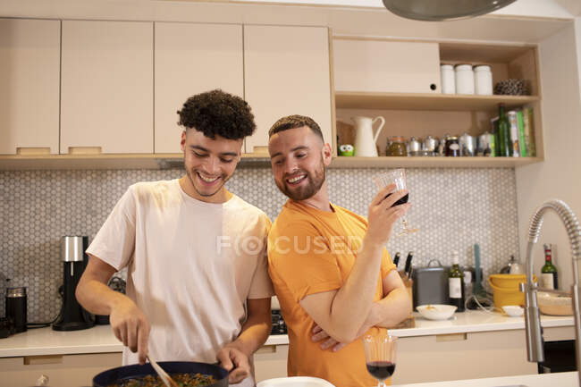 Feliz gay macho pareja cocinar y beber vino en cocina - foto de stock