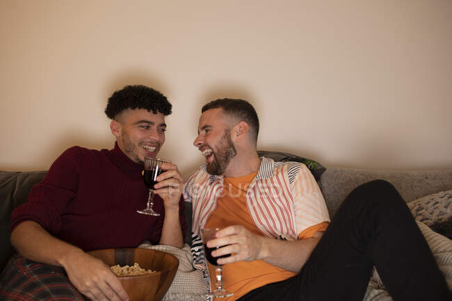 Feliz gay masculino casal beber tinto vinho e comer pipoca no casa — Fotografia de Stock