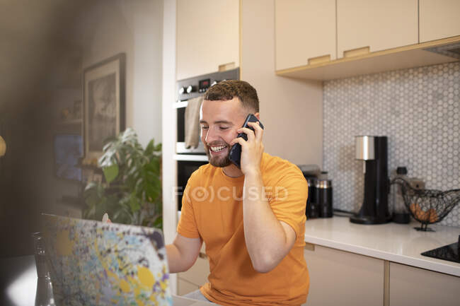 Счастливый молодой человек работает из дома разговаривает на смартфоне за ноутбуком — стоковое фото