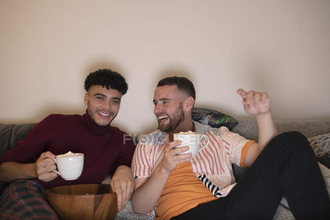 Happy gay male couple drinking hot cocoa watching TV on sofa — Stock Photo