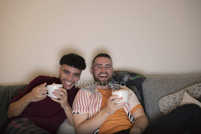 Happy gay male couple drinking hot cocoa and watching TV on sofa — Stock Photo